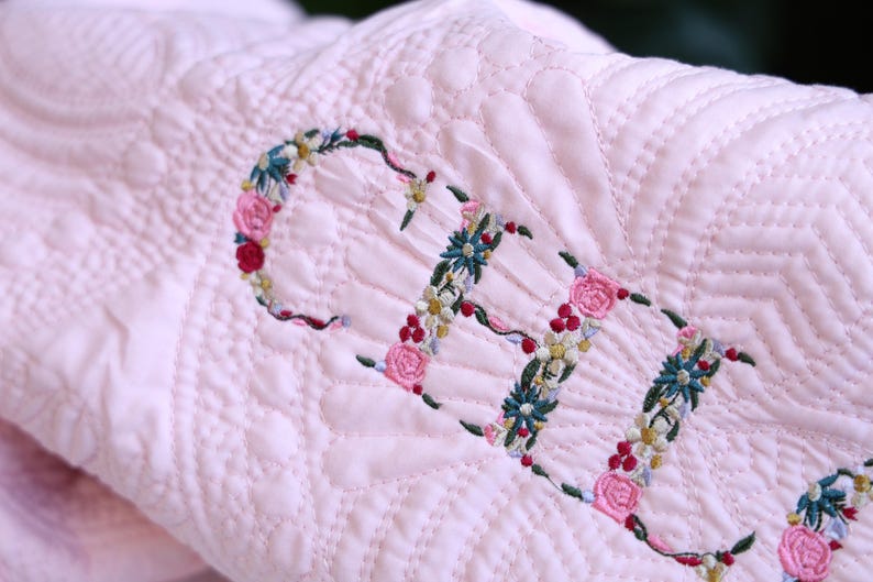 May include: A light pink quilted blanket featuring the name ELLE embroidered in a floral script. The letters are formed by intricate thread-stitched flowers in shades of pink, green, yellow, and blue, set against a textured, stitched fabric surface.