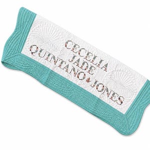 May include: A white quilted blanket with a teal border featuring a shell-like stitched pattern. The centre is embroidered with the text "CECELIA JADE QUINTANO JONES" in a floral-patterned font, with a small butterfly icon between the names.