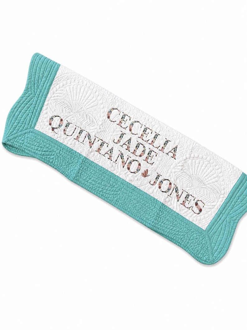 May include: A white quilted blanket with a teal border featuring a shell-like stitched pattern. The centre is embroidered with the text "CECELIA JADE QUINTANO JONES" in a floral-patterned font, with a small butterfly icon between the names.