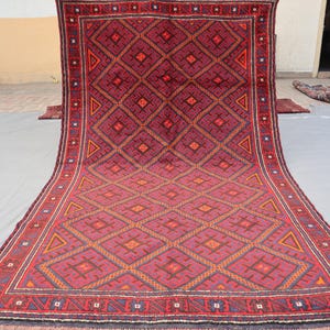 May include: A large, rectangular rug with a geometric pattern in shades of burgundy, red, and orange. The rug features a repeating diamond and triangle design, with a dark border and fringe along the edges. The rug is likely made of wool.