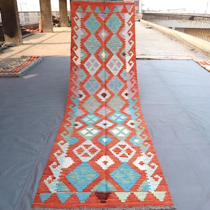 Könnte beinhalten: Ein langer, rechteckiger Teppich mit einem lebendigen geometrischen Muster. Der Teppich weist ein sich wiederholendes Rautenmuster in Orange-, Blau-, Weiß- und Grautönen auf. Der Teppich hat einen Fransenrand und ist wahrscheinlich aus gewebtem Material.