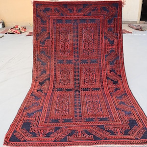 May include: A rectangular, hand-woven rug with a central design featuring geometric patterns in shades of red and navy blue. The rug's border has a repeating geometric design. The rug is displayed on a light gray surface.