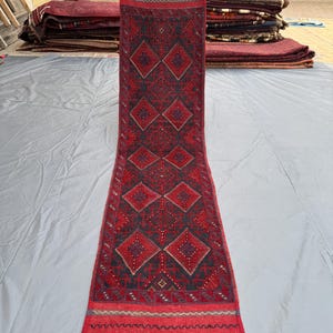 Może przedstawiać: Długi, prostokątny czerwony dywan z geometrycznym wzorem rombów w kolorze ciemnoczerwonym i czarnym. Dywan ma frędzel i jest trzymany przez osobę. Inne dywany są ułożone w stosy w tle.