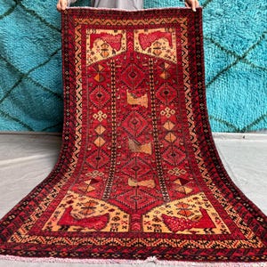 May include: A red rectangular rug with detailed geometric designs and bird motifs. The rug's central section has diamond shapes, and the borders have repeating patterns. The rug is shown against a blue backdrop.