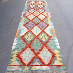 May include: A rectangular, colorful rug with a geometric diamond pattern. The rug features a red diamond grid with various colors including blue, green, yellow, and orange. The rug has a fringe on both ends.