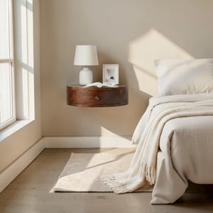 May include: A dark brown, floating nightstand with a single drawer, a white lamp, and a small framed picture. A book is open on the nightstand. The bed has a white comforter and a cream-colored throw blanket.