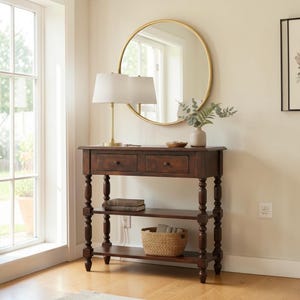 May include: A dark brown wooden console table with two drawers, two shelves, and turned legs. A gold-framed round mirror hangs above the table. A lamp with a white shade and a woven basket are on the table.