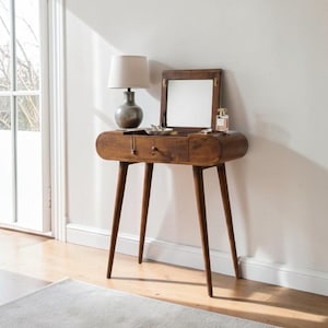 May include: A mid-century modern wooden vanity table with a flip-up mirror, a drawer, and slender, tapered legs. A small lamp and perfume bottle sit on top. The table is made of dark wood and is set against a white wall.