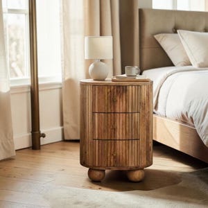 May include: A wooden nightstand with three drawers and a round top. The nightstand has a light-colored finish and is supported by three rounded feet. A small lamp with a white shade sits on top, along with a book and a mug. The nightstand is next to a bed.