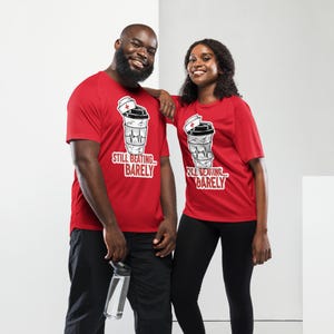Puede incluir: Dos personas vistiendo camisetas rojas con un gráfico de taza de café y el texto "Still Beating... Barely". El gráfico incluye un gorro de enfermera y una línea de latidos. Las camisetas son de manga corta.