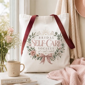 May include: A white canvas tote bag with dark red straps featuring a floral wreath graphic, a pink bow, and the text "BRIDAL SELF-CARE SOCIETY rest • reset • radiate". The design includes green leaves and small pink flowers surrounding the lettering.