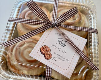 May include: A clear plastic container with a brown and white checkered ribbon tied around it. The container holds several cinnamon rolls with white frosting. A gift tag with the text "For the Rolls You Play! We appreciate you everyday!" is attached to the ribbon.