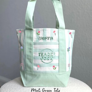 May include: A mint green and white striped canvas tote bag featuring an embroidered Trader Joe's logo and the name Sabrina in sage green thread. The bag is decorated with small pink and purple wildflower illustrations and has two sturdy handles.