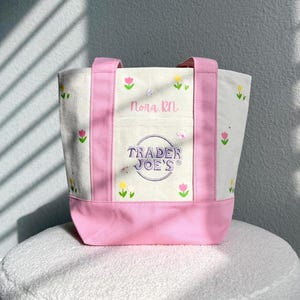 May include: A canvas tote bag with pink trim and handles. The bag displays the Trader Joe's logo and the name "Nora RN" embroidered on the front. Small embroidered flowers are scattered across the bag.