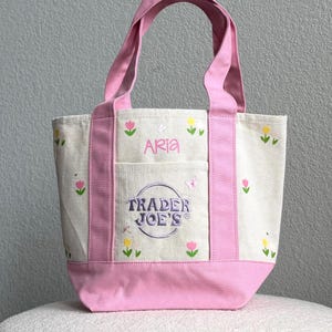 May include: A pink and cream canvas tote bag with embroidered flowers and the name "Aria". The bag features a front pocket and the "Trader Joe's" logo. The handles and base are a solid pink.