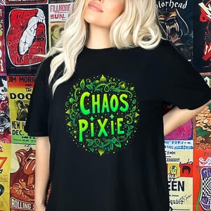 May include: Black t-shirt featuring a green and yellow design with the words "CHAOS PIXIE" in a circular pattern. The design is surrounded by leaves and small dots. The background is a collage of concert posters.