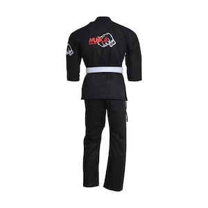 Puede incluir: Uniforme negro de artes marciales con cinturón blanco. El uniforme tiene el texto "MUKKA Sports" y un gráfico de puño en rojo y blanco en la espalda y la manga izquierda. El uniforme incluye chaqueta y pantalón.