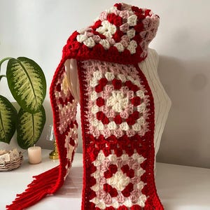 Könnte beinhalten: Handgefertigter Häkelschal im Granny-Square-Muster. Der Schal hat einen roten Rand und Fransen mit roten, weißen und rosa Quadraten. Der Schal wird auf einer weißen Oberfläche präsentiert.