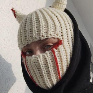 Puede incluir: Pasamontañas de punto color crema con dos cuernos pequeños y detalles rojos. El pasamontañas tiene detalles de hilo rojo alrededor de los ojos y en las mejillas. La persona lleva una sudadera con capucha negra.