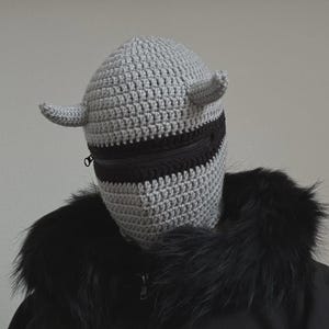 May include: A gray crocheted balaclava with two small horns and a black zippered section across the face. The balaclava is worn over a black jacket with a fur collar. The design is unique and could be used for costume or fashion.