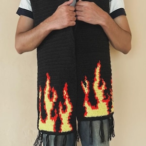 Puede incluir: Bufanda negra de ganchillo con un diseño de llamas amarillas y rojas. La bufanda tiene flecos en la parte inferior. Las llamas están delineadas en rojo y rellenas de amarillo. La bufanda se lleva sobre una camisa blanca y negra.