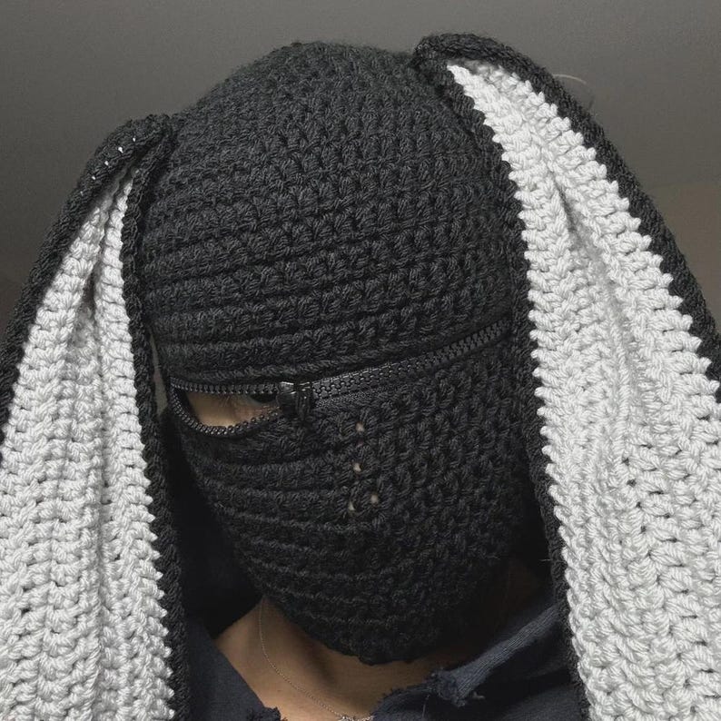 May include: A black and gray crochet balaclava with bunny ears. The balaclava features a zipper across the face and is made of a textured, handmade yarn. The ears are lined with white yarn.