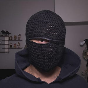 以下が含まれることがあります： 目のためのジッパー開口部が付いた黒色の編み物バラクラバ。ヘッドカバーは厚く、質感のある糸でできており、濃い青色のフード付きスウェットシャツの上に着用されています。