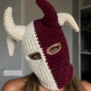 May include: A two-toned balaclava crafted from plush, velvety yarn, featuring a split design in cream and deep burgundy. The headpiece includes three prominent, curved, horn-like protrusions extending from the top and sides, with eye openings.
