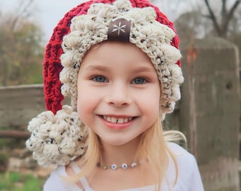 May include: A red and cream-colored crocheted hat with a pom-pom and a brown leather tag with a star design. The hat is worn by a young person with blonde hair and a white shirt.