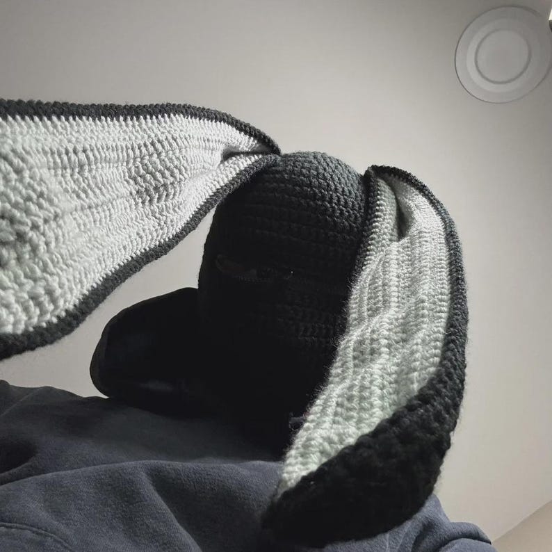 May include: A black crochet balaclava with long bunny ears. The ears are black with a light gray interior. The hat is made of a soft, textured yarn, and the design is a unique and playful accessory.