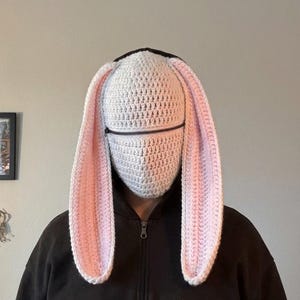 May include: A white crocheted bunny hood with long, pink-lined ears and a black zipper across the face. The hood is worn over a black hooded sweatshirt, creating a whimsical and playful look. The ears are long and floppy.