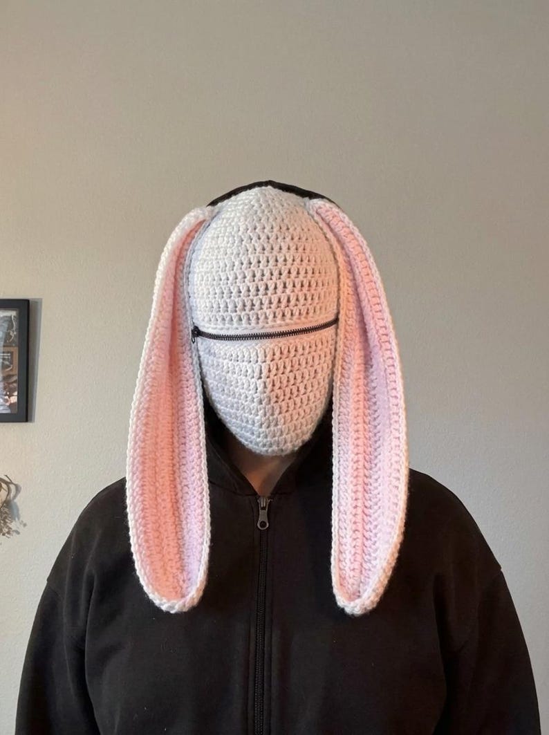 May include: A white crocheted bunny hood with long, pink-lined ears and a black zipper across the face. The hood is worn over a black hooded sweatshirt, creating a whimsical and playful look. The ears are long and floppy.