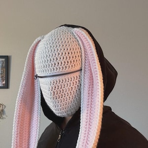 May include: A white crochet bunny mask with long, floppy ears, lined with pink. The mask features a zipper across the mouth area and is worn over a black hooded sweatshirt. The handmade mask is a unique costume accessory.