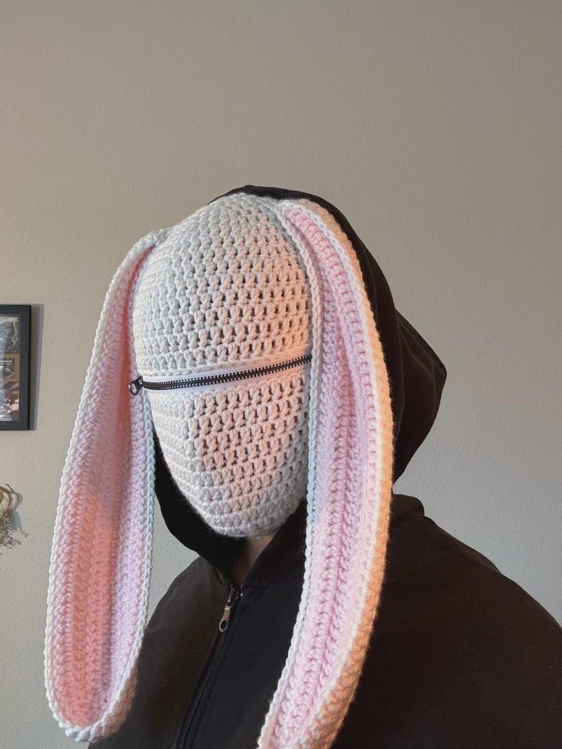 May include: A white crochet bunny mask with long, floppy ears, lined with pink. The mask features a zipper across the mouth area and is worn over a black hooded sweatshirt. The handmade mask is a unique costume accessory.
