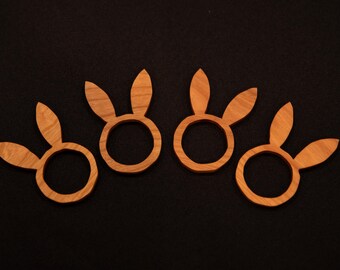 Puede incluir: Cuatro servilleteros de madera con forma de conejito. Cada anillo tiene un centro circular con dos orejas verticales. La madera es de color marrón claro y tiene una veta visible. Los servilleteros están colocados sobre un fondo oscuro.