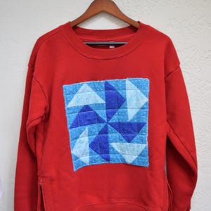May include: A red sweatshirt with a square, patchwork design on the front. The patchwork features a pinwheel pattern in shades of blue and white. The sweatshirt has a zipper detail on the side.
