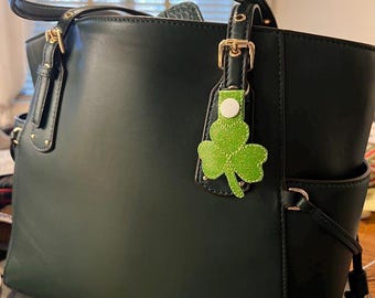 May include: Dark green tote bag with a matching handle and gold-colored hardware. A green glitter shamrock charm hangs from the handle. The bag has a structured shape with a side pocket.