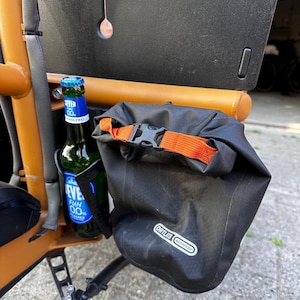 Könnte beinhalten: Eine schwarze, wasserdichte Ortlieb-Rolltop-Tasche mit orangefarbenem Riemen und Schnalle an einem orangefarbenen Fahrradrahmen. Neben der Tasche befindet sich eine grüne Glasflasche Jever Fun 0.0 Bier in einem schwarzen Flaschenhalter.
