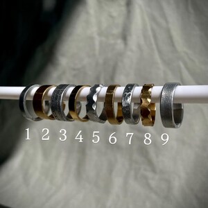 Può includere: Una collezione di nove anelli con vari design e finiture, esposti su un'asta bianca. Gli anelli presentano diverse trame e colori, tra cui argento, oro e tonalità metalliche scure. I numeri da 1 a 9 sono stampati sotto.