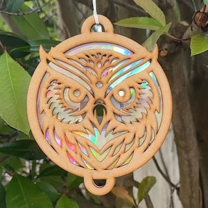 May include: Wooden owl ornament with intricate cut-out details. The owl's face features a holographic, iridescent insert that reflects multiple colors. The ornament is suspended by a white string, perfect for hanging. The design is circular with a decorative top and bottom.