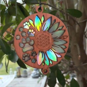 May include: Wooden round ornament featuring a sunflower design with iridescent petals and paw prints. The ornament is brown with a hole for hanging. The sunflower petals have a rainbow sheen.