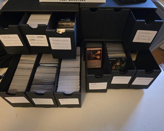 May include: A black storage box with multiple drawers filled with trading cards. The drawers are labeled with handwritten text, including "Lands", "Tokens", "Foils", "Commander", "Modern", "Multicolored", "Red", "Blue", and "Black".