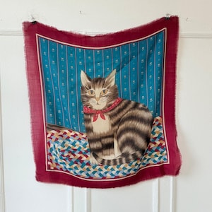 Puede incluir: Un pañuelo cuadrado con una ilustración de gato. El pañuelo tiene un borde burdeos y un centro azul turquesa con un estampado de rayas. El gato es marrón y gris con un collar rojo. El pañuelo está colgado en una puerta blanca.