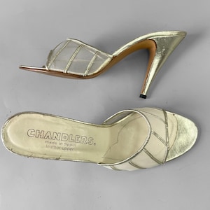 May include: A pair of vintage gold-colored high-heeled sandals. The sandals feature a sheer mesh upper with gold accents and a pointed toe. The heels are a metallic gold color. The sole of one sandal is visible, with the brand name "CHANDLERS" printed on it.