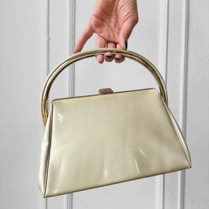 May include: A vintage cream-colored handbag with a glossy finish. It features a gold-toned, arched handle and a rectangular clasp. The bag's shape is trapezoidal, with a structured design.