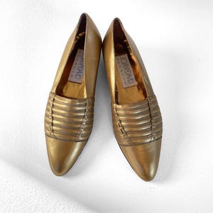 May include: A pair of gold-colored leather flats. The shoes have a pointed toe and a decorative horizontal stitched design across the top. The brand name "JOAN & DAVID" is visible inside the shoe.