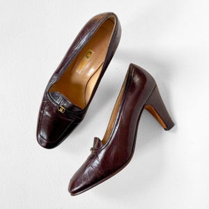 Peut inclure: Une paire d'escarpins en cuir marron foncé à petit talon. Les chaussures ont un bout carré et un détail doré sur le dessus. L'intérieur de la chaussure est de couleur beige clair, et le nom de la marque est visible.