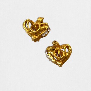 May include: Two gold-colored heart-shaped earrings. Each earring features a textured design with a small, clear gemstone embellishment. The earrings have a vintage aesthetic and are set against a white background.