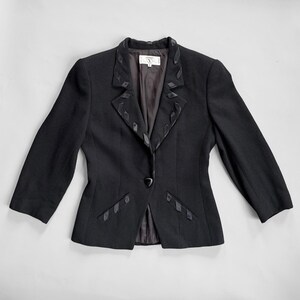 May include: Black blazer with decorative ribbon detailing. The jacket has a single-button closure and a notched lapel with ribbon accents. The sleeves are three-quarter length. A small label is visible inside the blazer.
