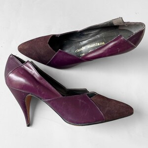 May include: A pair of purple suede and leather high heel pumps with a pointed toe. The shoes have a cut-out design and the label "claude montana" is visible on the inside of one shoe.